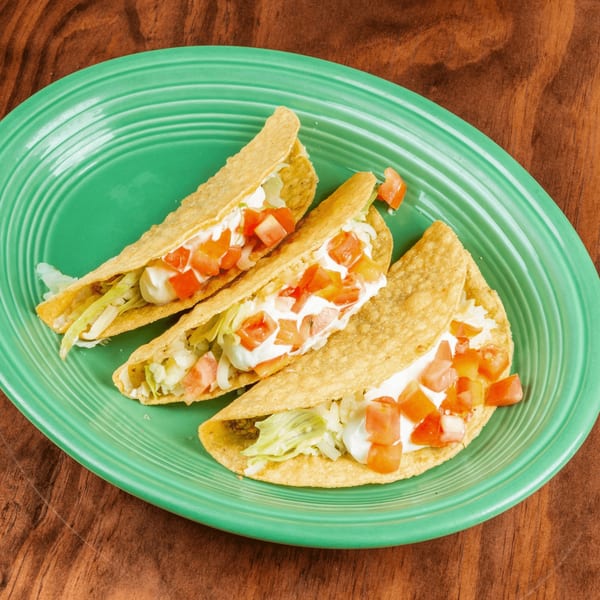 Three crispy tacos filled with meat, lettuce, sour cream, and diced tomatoes on a green plate.
