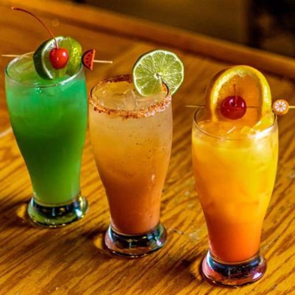 Three colorful Mi Casa Mexican Restaurant cocktails, garnished with fruit, on a wooden bar.